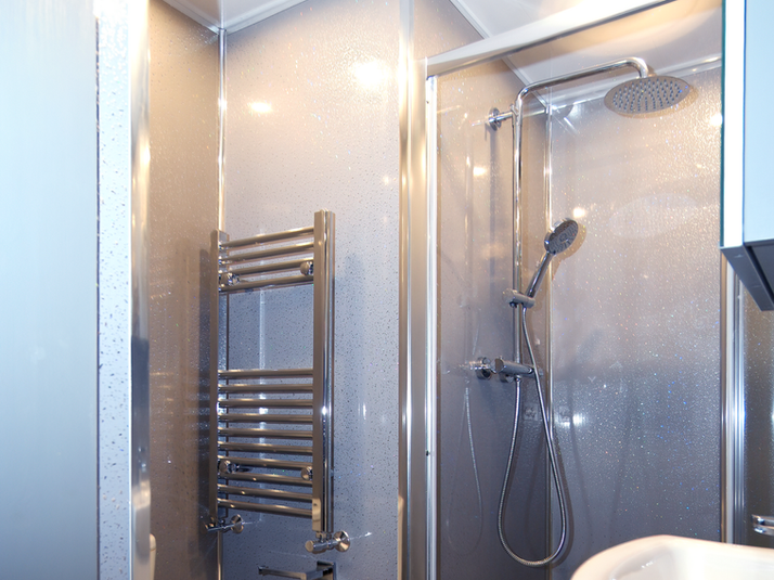 A modern en suite shower room with a silver panelled shower enclosure, chrome rainfall and handheld shower heads, and a compact white vanity sink. The design is bright and clean with reflective surfaces and grey paneling on the adjacent wall.