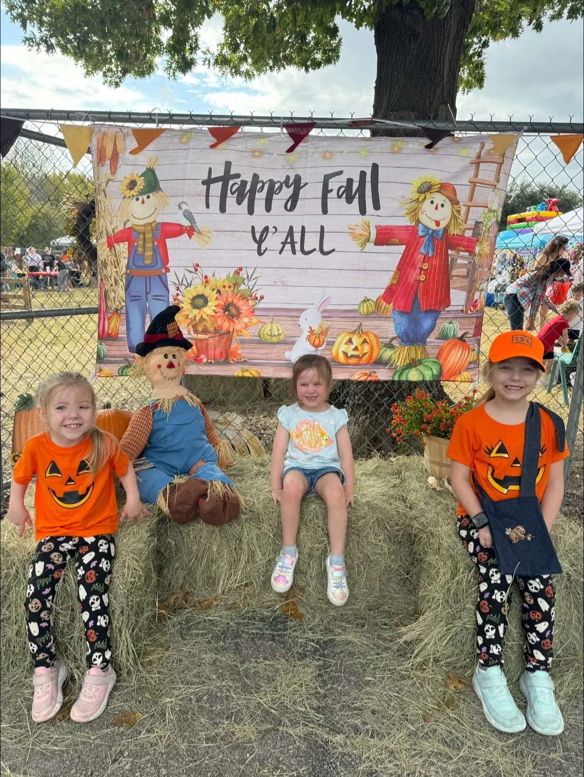 Three smiling children beside hay bales and a Happy Fall Y’all display at the school fall festival