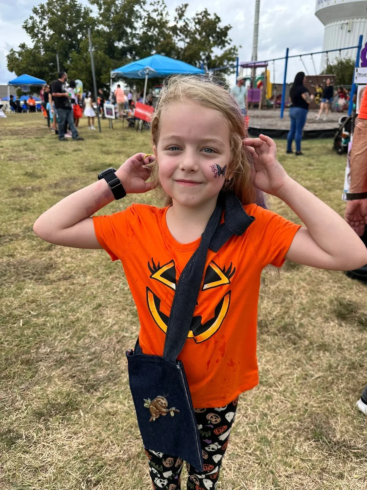 A child in an orange pumpkin shirt showing face paint at the outdoor fall festival