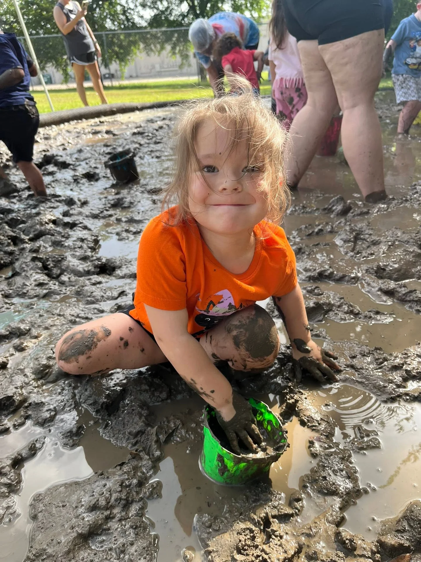 A smiling child filling a green bucket with mud during outdoor play at The Children’s Spot