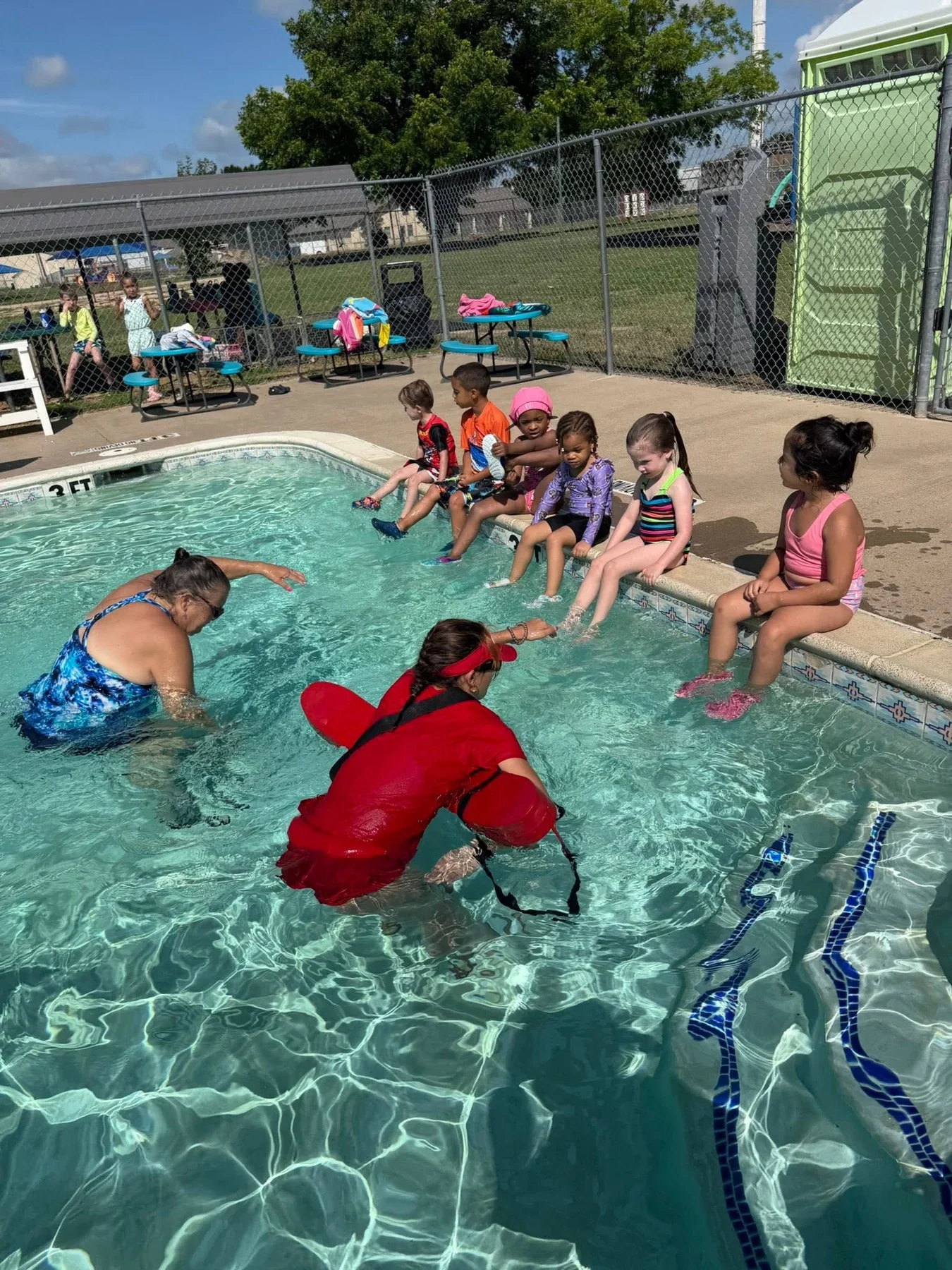 Children sitting along the school pool with two adults in the water during a summer activity