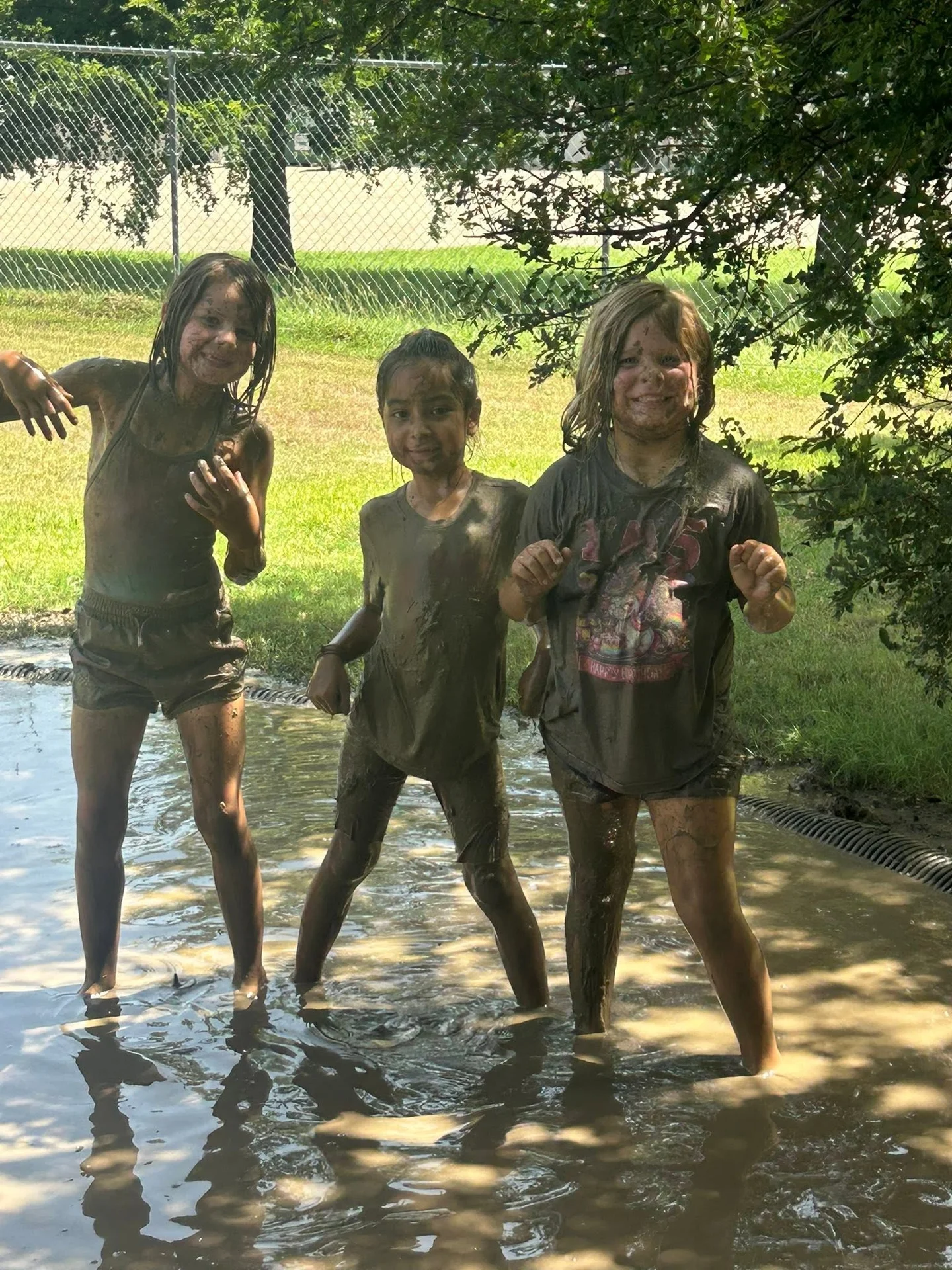 Three children smiling together, covered in mud during the school’s Mud Day in June 2026