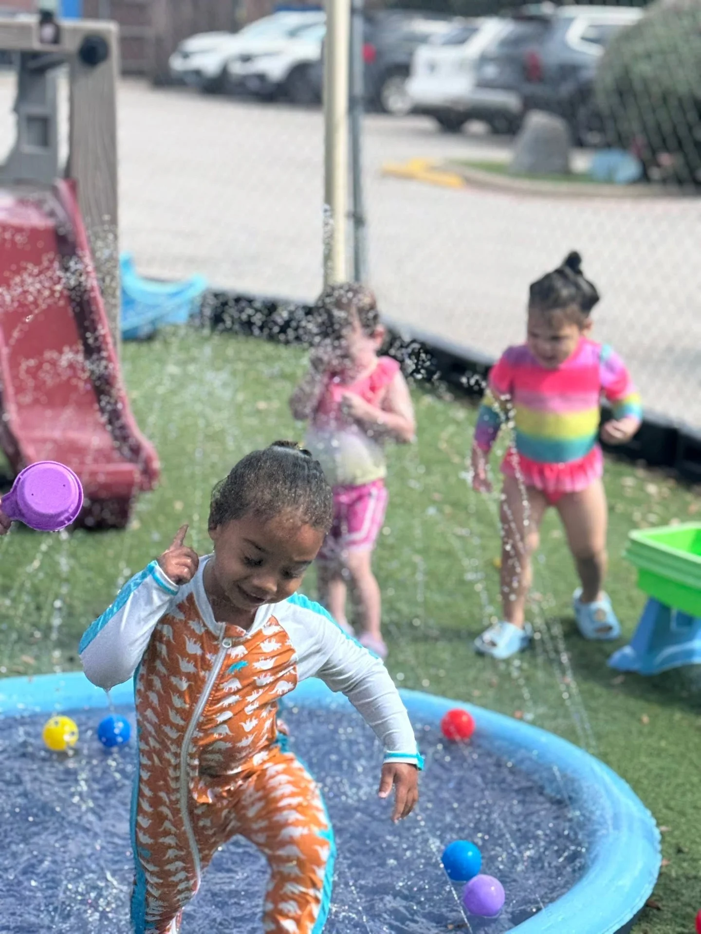 A smiling child stepping through sprinklers with friends in the school’s outdoor play area
