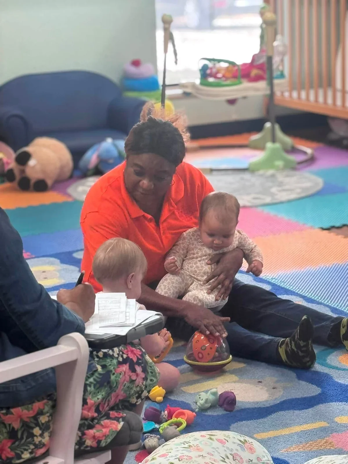 A caregiver sitting on the classroom floor, holding a baby and playing with another young child
