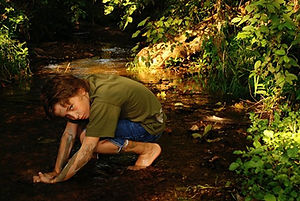 A little boy is crouched in the creek and covered in mud staring intensely into the camera.