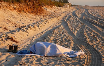 A covered figure sleeps on the beach with their boots lying next to them