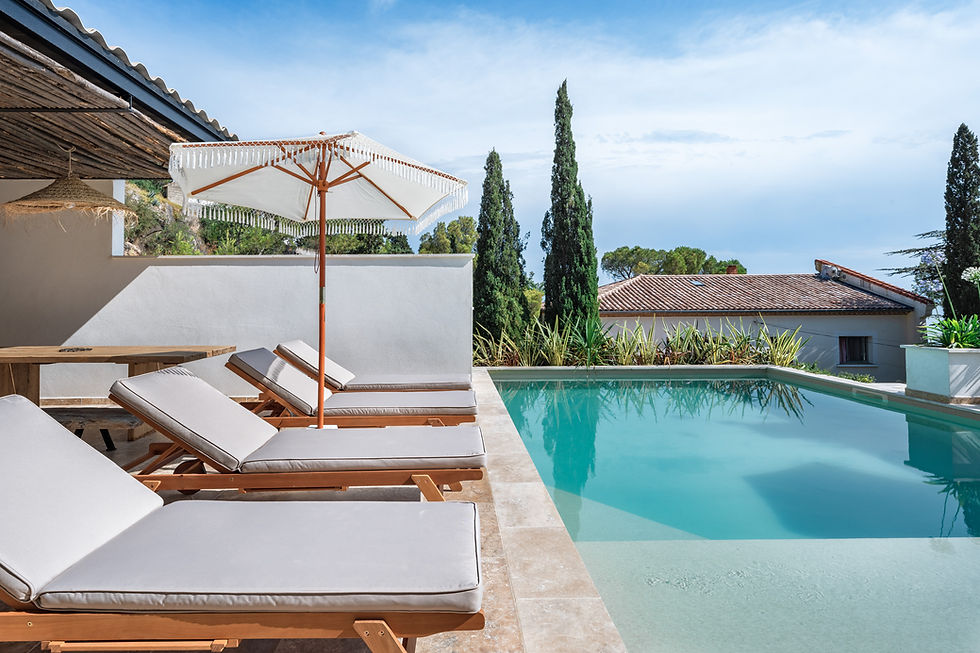 Chaises longues sous un parasol près d'une piscine turquoise. Fond de cyprès et maison au toit rouge. Atmosphère sereine et ensoleillée.