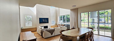 Spacious, sunlit living room with high ceilings, featuring a wooden dining table, cozy sofa, artwork on wall, and large windows showing a garden view.