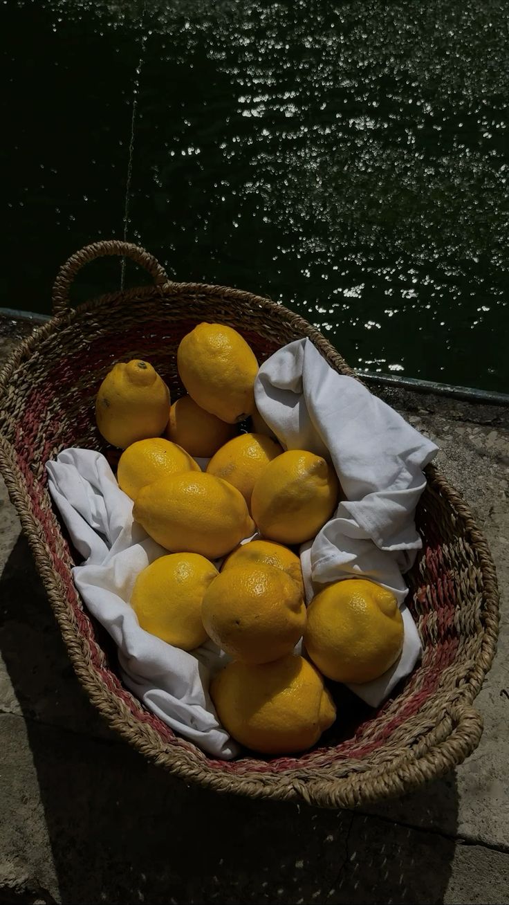 Ripe yellow lemons in a woven basket with a white cloth beside the water.