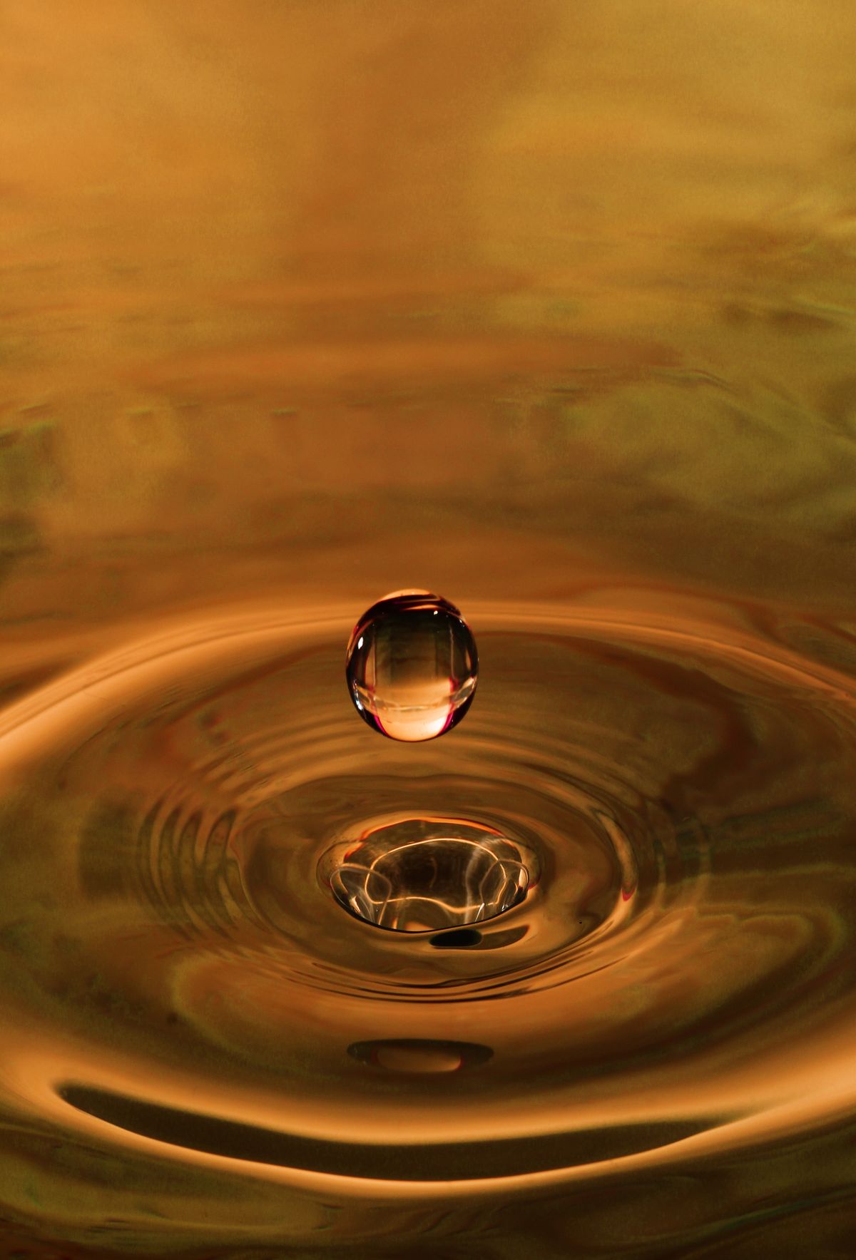 Ein einzelner Wassertropfen über einer goldfarbenen Wasseroberfläche mit kreisförmigen Wellen.