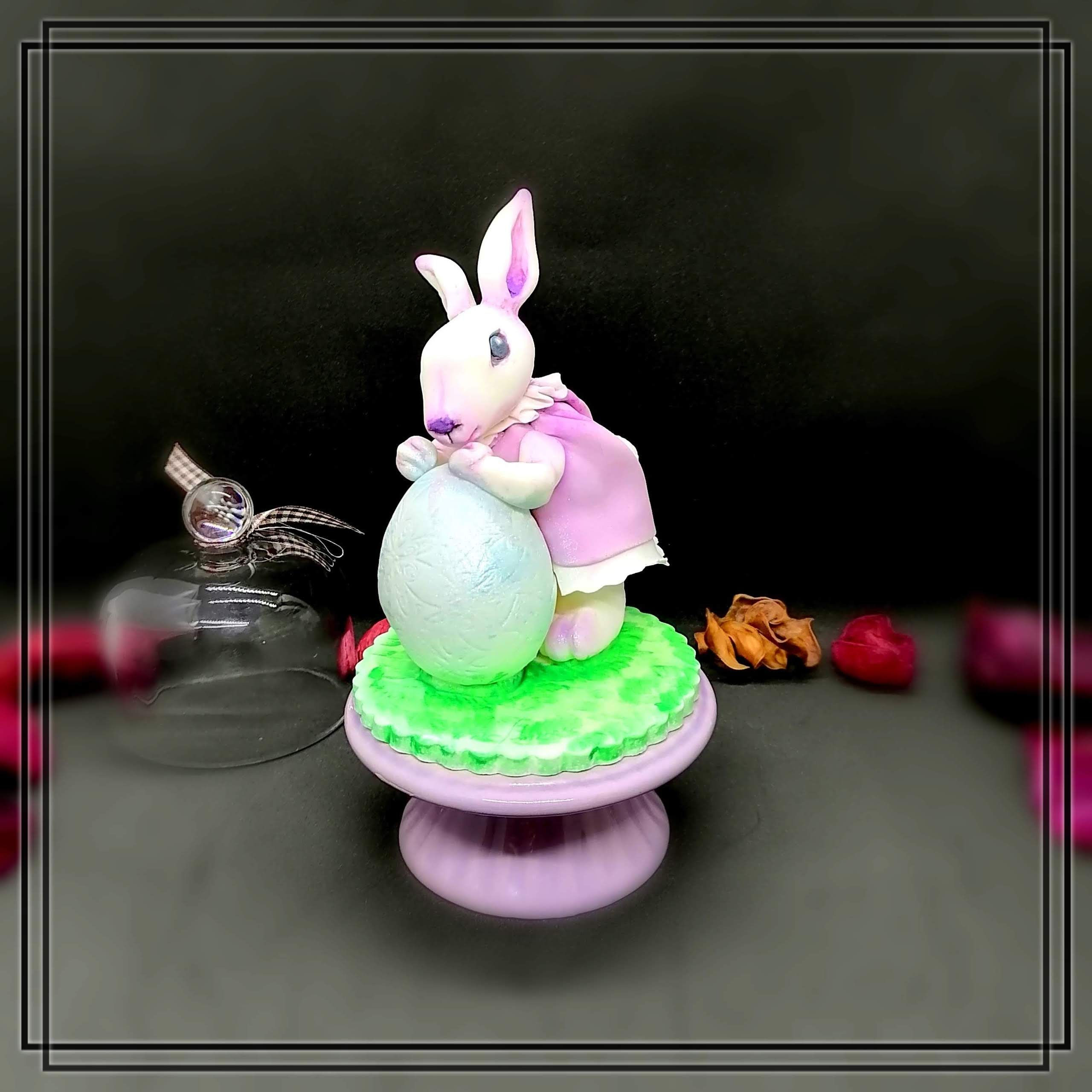Handmodellierter Osterhase aus Fondant mit rosa Kleid und hellblauem Osterei auf grüner Wiese, essbare Tortendeko für Ostern