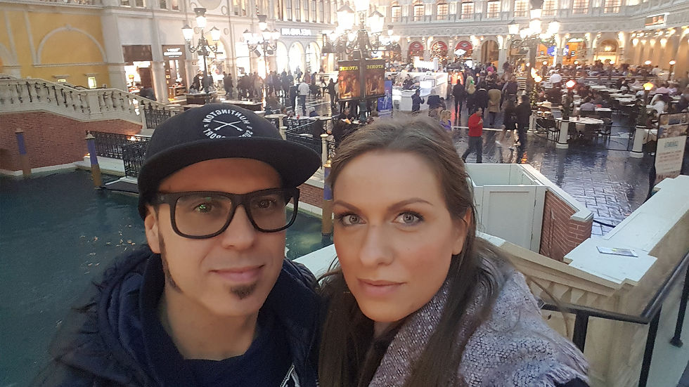 A couple poses indoors, surrounded by a busy shopping area at the Venetian in Las Vegas with elegant architecture and warm lighting. The atmosphere is lively and festive.