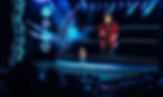 Shin Lim Magician on stage in Las Vegas in red jacket performing for an audience. Neon playing card symbols and blue lights create a dramatic atmosphere.