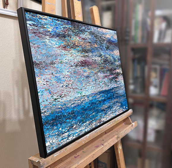 Side view of oil painting "The Rough Sea" by Catherine Snowstorm in its black metal frame, on the studio easel. The angled perspective reveals the thick palette knife impasto surface — layers of paint building up visibly from the canvas edge. The vivid turquoise sea is at the lower right. Palette knife seascape, alla prima, impasto oil painting, turquoise and navy.