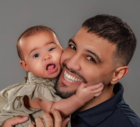  Pai segurando seu bebê, ambos sorrindo, capturando um momento de alegria e ternura em um fundo cinza neutro.
