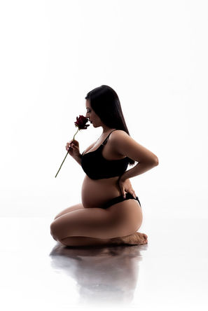 Ensaio fotográfico de gestante no estúdio, com a mãe ajoelhada em um fundo branco, vestindo lingerie preta enquanto segura uma rosa vermelha perto do rosto. A imagem minimalista destaca a serenidade e a conexão da mãe com a maternidade."