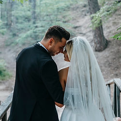 A couple in wedding attire stands on a wooden bridge amidst a serene forest setting.