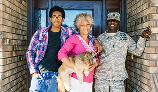 A family of three stand together. Two grown sons stand on either side of their mother who is holding a dog. The son to the left has his hand in his pocket and the other around his mother. The son to the right is in the US army uniform and is leaning against the nearby wall. All three people are wearing relaxed smiles.