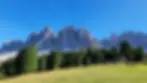 Mountain peaks under a clear blue sky, with a forest at the base and a grassy field with a playground in the foreground. Italian Dolomites