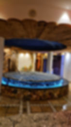 Circular hot tub with bubbling water, stone base, and blue lighting. Cozy spa room with wooden beams and mural on the wall. Orange towel hanging.