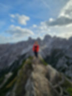 Woman in a red jacket stands on a rocky mountain peak with dramatic jagged cliffs and a blue sky with clouds in the background. Cadini Di Misurina Viewpointin the Italian Dolomites