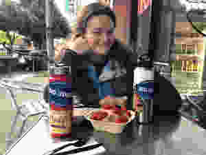 Tess smiles at the camera. She has white skin, has her long dark blonde hair fall on her shoulders. In front of Tess is a paper box with a cinnamon bun with raspberries on top. There are two water bottles with Big Ideas stickers on either side of the cinnamon bun. In the background is the pavement, trees and the inside of Cinnaholic Bakery can be seen.