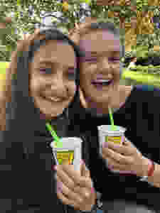 Yagmur and Tess smile at the camera, holding white paper cups that have Yogurt Park written on them in green and there is a florescent green plastic spoon in them. Yagmur has long brown hair, she is fair skinned, wears a dark green scarf and dark green top. Tess has a big smile on her face, has white skin and her hair in a bun. She wears a dark blue top. In the background is a grassy area and a big tree with many branches and green leaves.