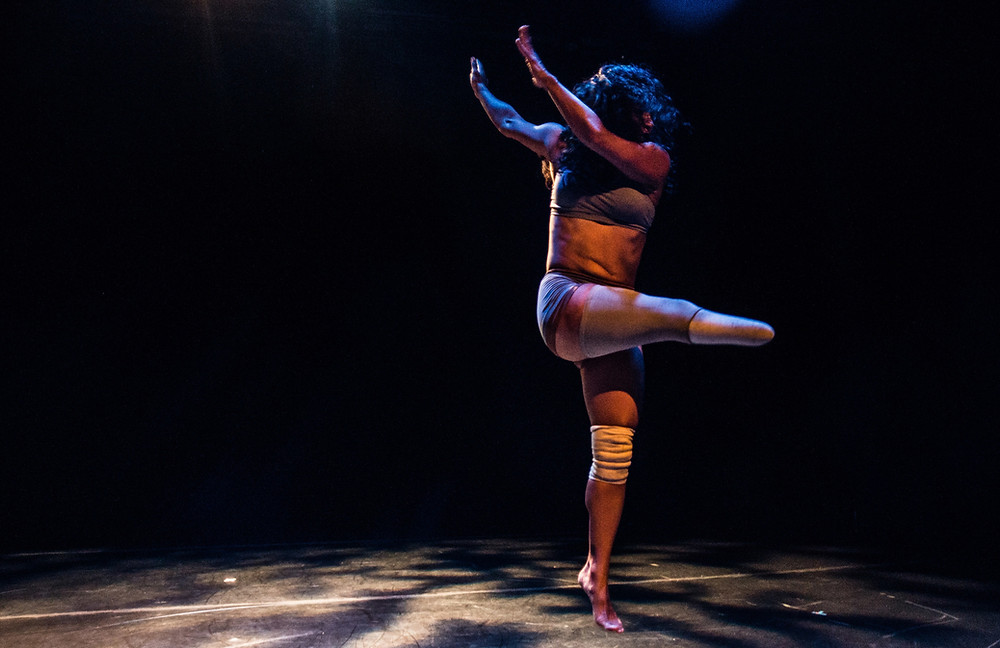 Stephanie holds a pose with her arms lifted up to her right side, her head twisted to the left as she stands on her left toes and points her right lower limb to her left side. The background is all dark, but there is a patterned light shining on her as she holds the pose. She has dark black hair that covers her face and she is wearing a purple pastel colored crop top and shorts, as well as a white knee brace on her left knee. She wears a limb socket for her right lower limb.