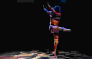 Stephanie holds a pose with her arms lifted up to her right side, her head twisted to the left as she stands on her left toes and points her right lower limb to her left side. The background is all dark, but there is a patterned light shining on her as she holds the pose. She has dark black hair that covers her face and she is wearing a purple pastel colored crop top and shorts, as well as a white knee brace on her left knee. She wears a limb socket for her right lower limb.