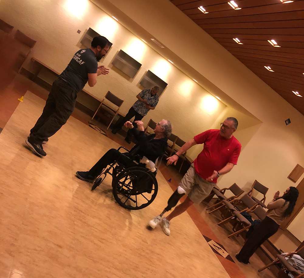 Three participants at amputee support group pictured together as they are dancing. One of the participants is in a wheelchair, and two other participants are standing. One of the participants on the right has their prosthetic for their right lower limb exposed.