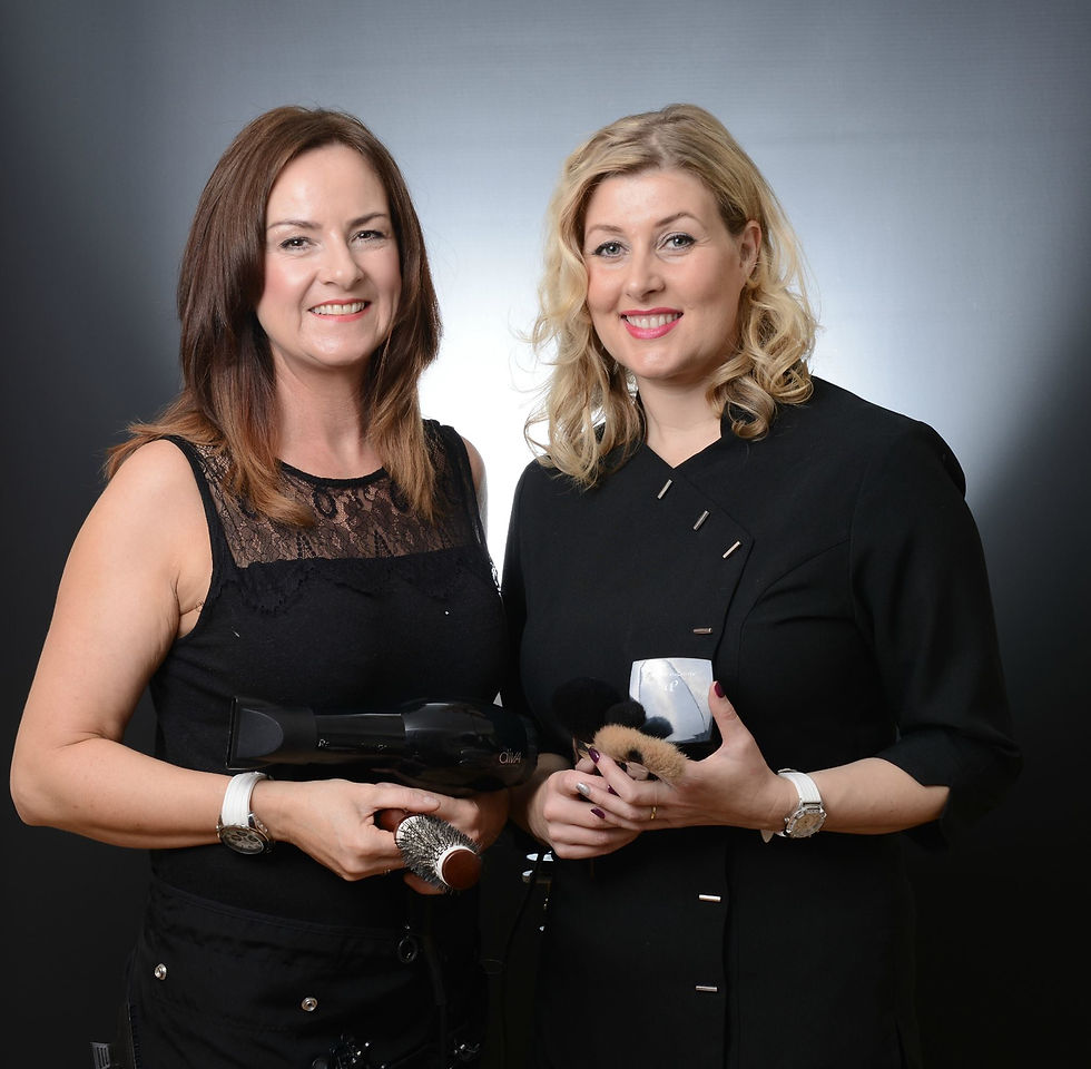 Two women smiling, holding hairdressing tools in a studio setting. Both wear black outfits; one holds a hair dryer, the other, brushes.