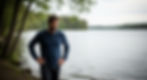 Man in a blue sweater stands with hands on hips by a calm lakeside, surrounded by trees. The scene is serene and introspective.