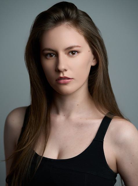 Victoria Nelson with long straight brown hair poses against a neutral gray background, wearing a sleeveless black top. Victoria has a serious facial expression, natural makeup, and an even complexion. The image is a high-resolution studio portrait, ideal for professional or modeling portfolio use.