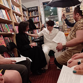 Various people sit in a bookshop. One of them is singing.