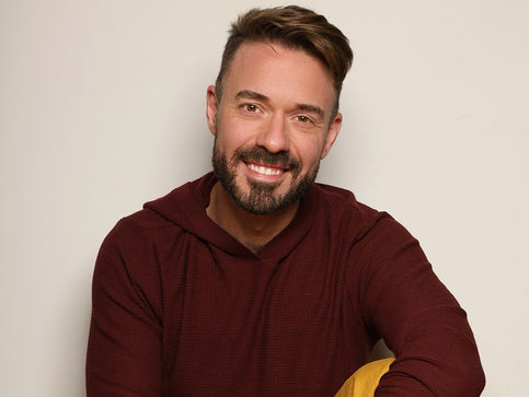 Foto do ator com blusa vermelha e calça caqui, sentado no chão, abraçando a perna esquerda, dando um sorriso simpático