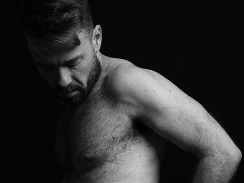 foto artística do ator de pé, de lado, sem camisa, olhando para baixo com as mãos por dentro da cueca pela parte de trás de seu corpo. 