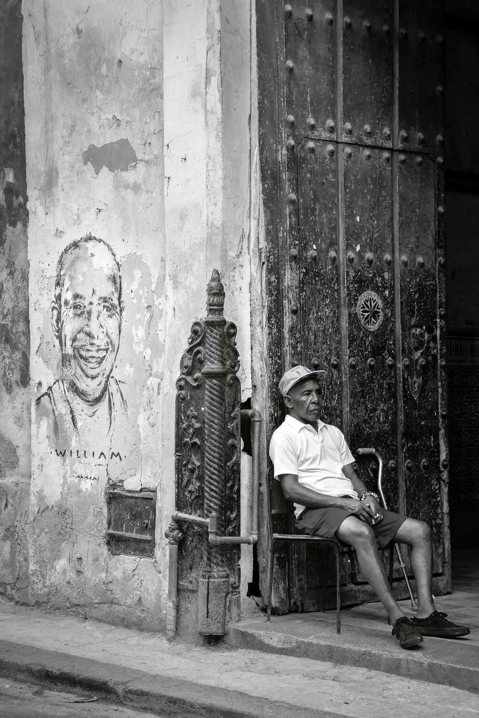 Black and white street photograph of a Cuban man sitting near a large wooden door beside a painted mural on a weathered wall in Havana.