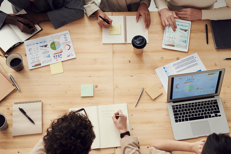 On a pine table there are several people sitting at the table. Two have open notebooks ready to write in. Another person towards the back has a tablet with graphs on it. On the left corner a person has a printed out page of graphs. And in the right lower corner, a person has a laptop open to a PDF with charts.