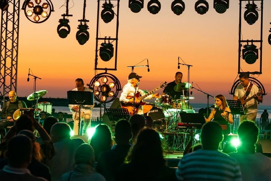 Fawzi Chekili sur la scène de "Street Jazz" le 6 juillet 2026 à Tabarka. Outdoor band performs at sunset by the sea, with audience silhouettes and green stage lights.