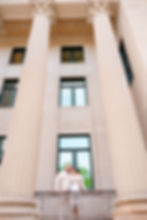 Bride and groom embrace on a balcony between towering cream columns, with dark windows and a calm, elegant mood. . Mecklenburg County Courthouse wedding by Aubrey Elizabeth Photography and Charlotte NODA wedding photos.