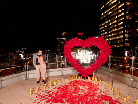 Couple kissing on a rooftop at night, surrounded by rose petals and candles. A "Will You Marry Me?" sign framed by a heart of red roses. Romantic mood. Charlotte private patio skyline proposal by Aubrey Elizabeth Photography.