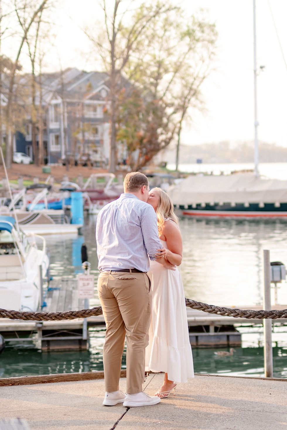 A couple kisses by a marina under trees on a sunny day. Boats and houses in the background. A sign reads "PRIVATE SLIP." Romantic mood. A man kneels proposing to a smiling woman in a white dress by a marina with boats. Trees and buildings in the background create a serene mood. Lake Norman Proposal at North Harbor Club.