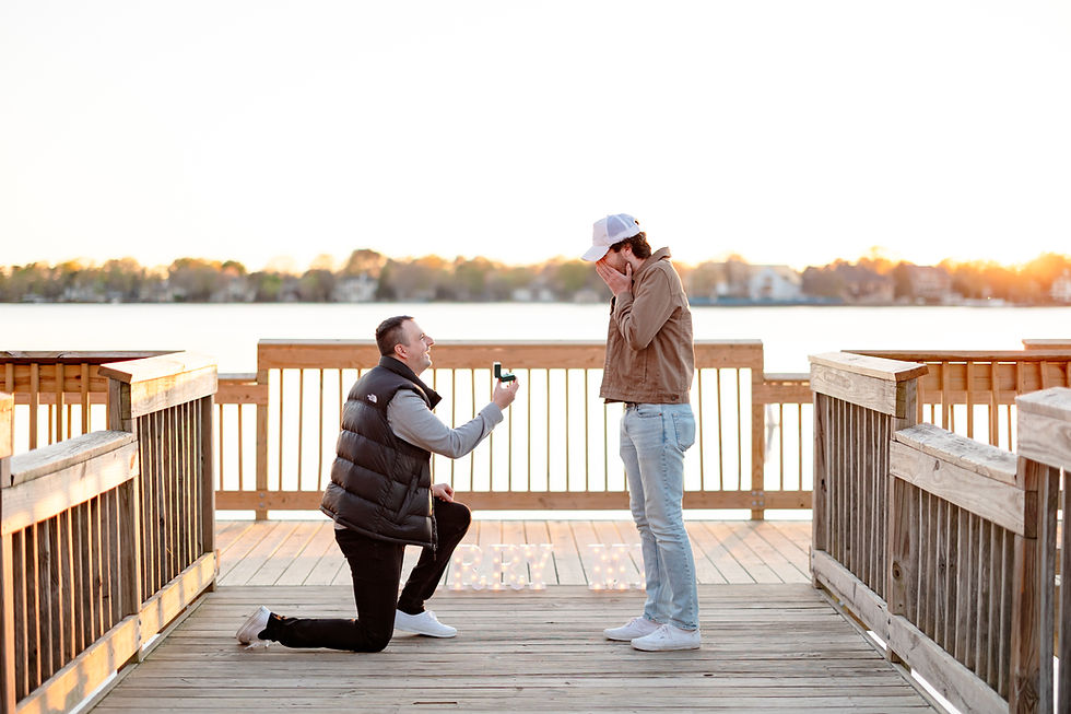 Man kneels proposing on a boardwalk at sunset, holding a ring. Another man stands surprised, hands over mouth. "Marry Me" lit in background.