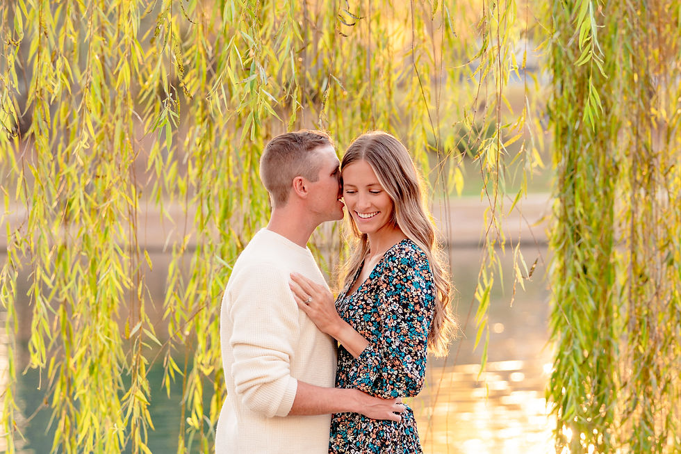 Couple embracing under willow branches near a serene lake at sunset. The man whispers to smiling woman in a floral dress. Romantic mood. Freedom Park, Charlotte, North Carolina.