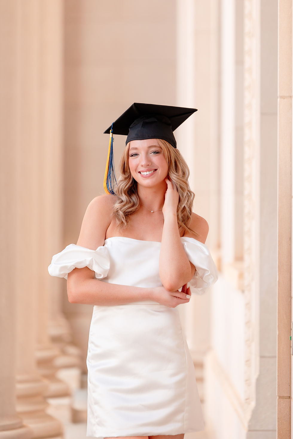 Young woman in white dress and graduation cap smiles confidently, standing in a sunlit hallway with light stone columns. A woman in a white dress holds a graduation cap on a rooftop with blurred buildings in the background, during sunset. Senior cap and gown session in Charlotte, North Carolina.