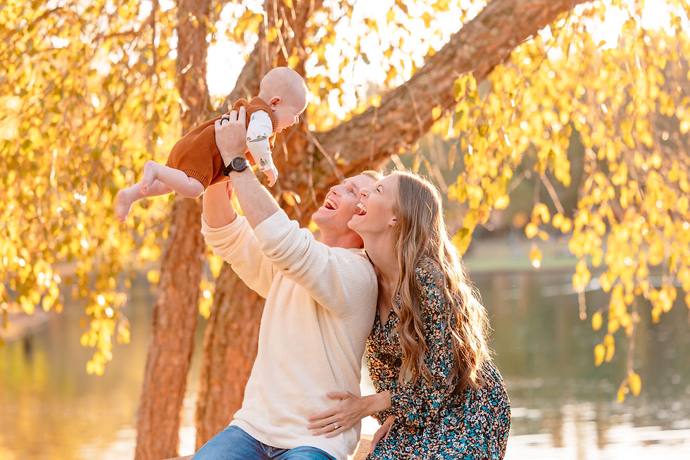 A couple joyfully lifts a baby under a tree with golden leaves by a lake. Warm sunlight creates a happy, serene atmosphere. Freedom Park, Charlotte, North Carolina.