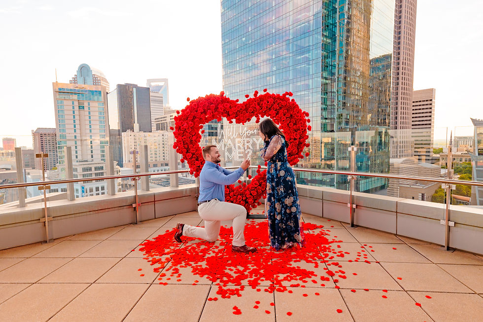 Man proposing on rooftop with city skyline. Red heart of roses, "Will you marry me?" text. Romantic, joyful atmosphere. Proposal in uptown Charlotte with city skyline. 8ft Marry Me lights and rose arch. rose petal decor.