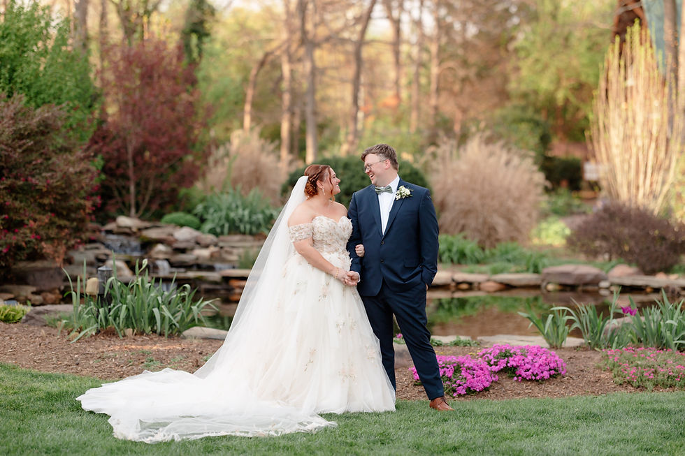 Bride in white gown and groom in navy suit smiling at each other in a garden setting with lush greenery and pink flowers. Wedding at Alexander Homestead in Charlotte, NC by Aubrey Elizabeth Photography
