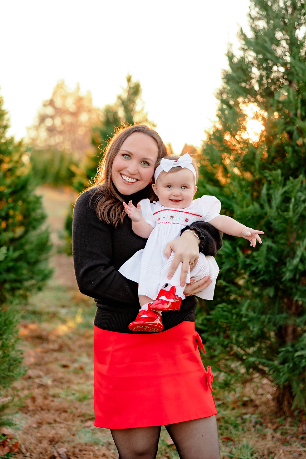 Smiling woman holds a cheerful baby in a white dress with red shoes, set in a sunlit evergreen forest. Mood is joyful and bright.