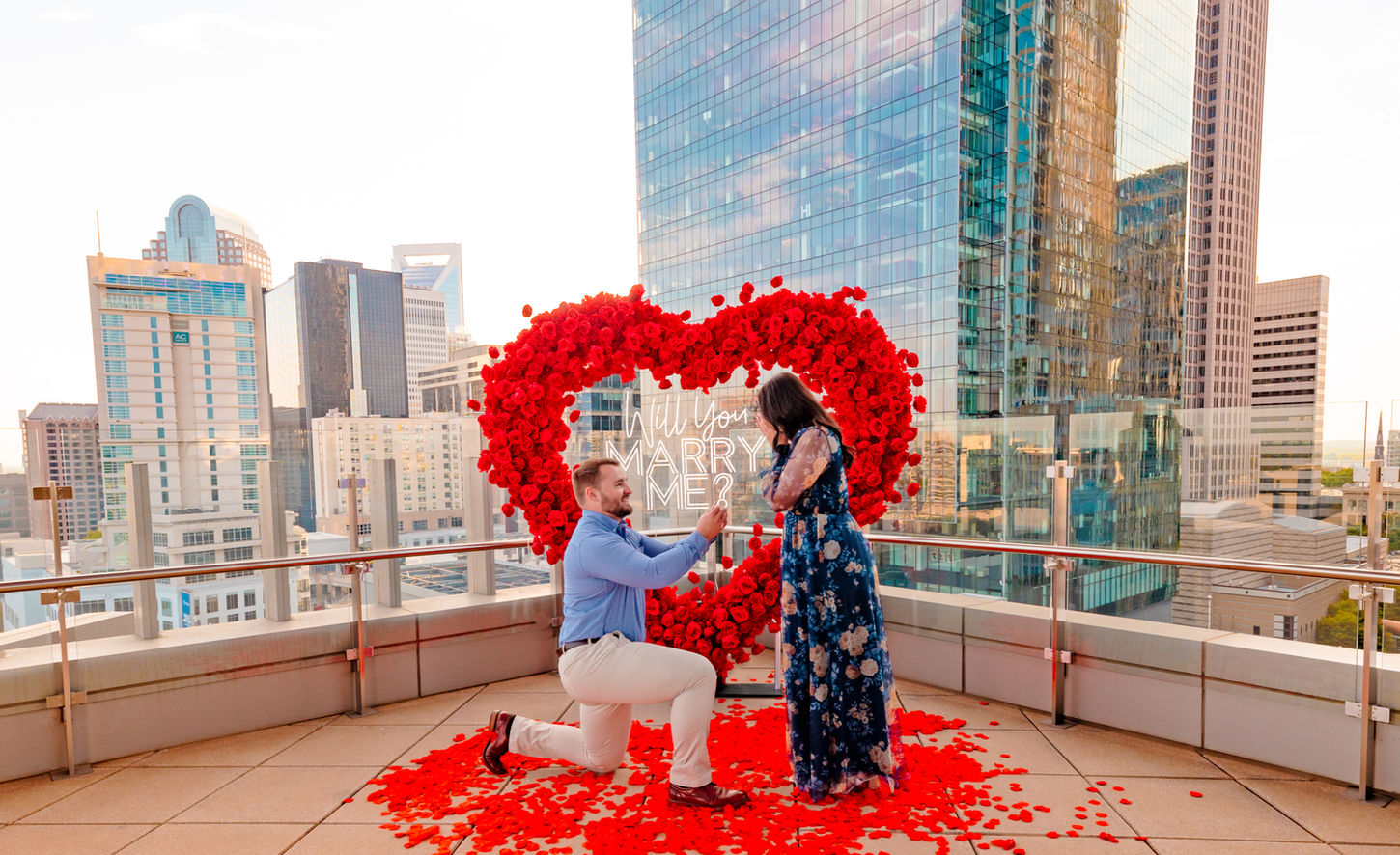 Luxury rooftop marriage proposal overlooking the Charlotte NC skyline with white floral arch, candles, rose petals and proposal photography